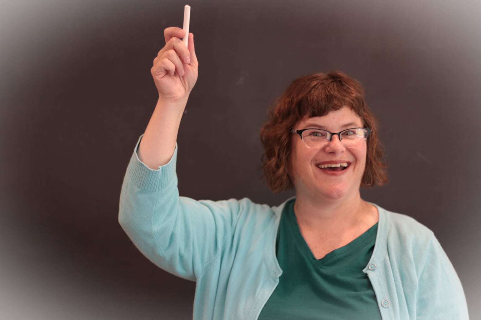 woman in a teal shirt and aqua sweater in front of a slate board with her arm raised holding a piece of chalk