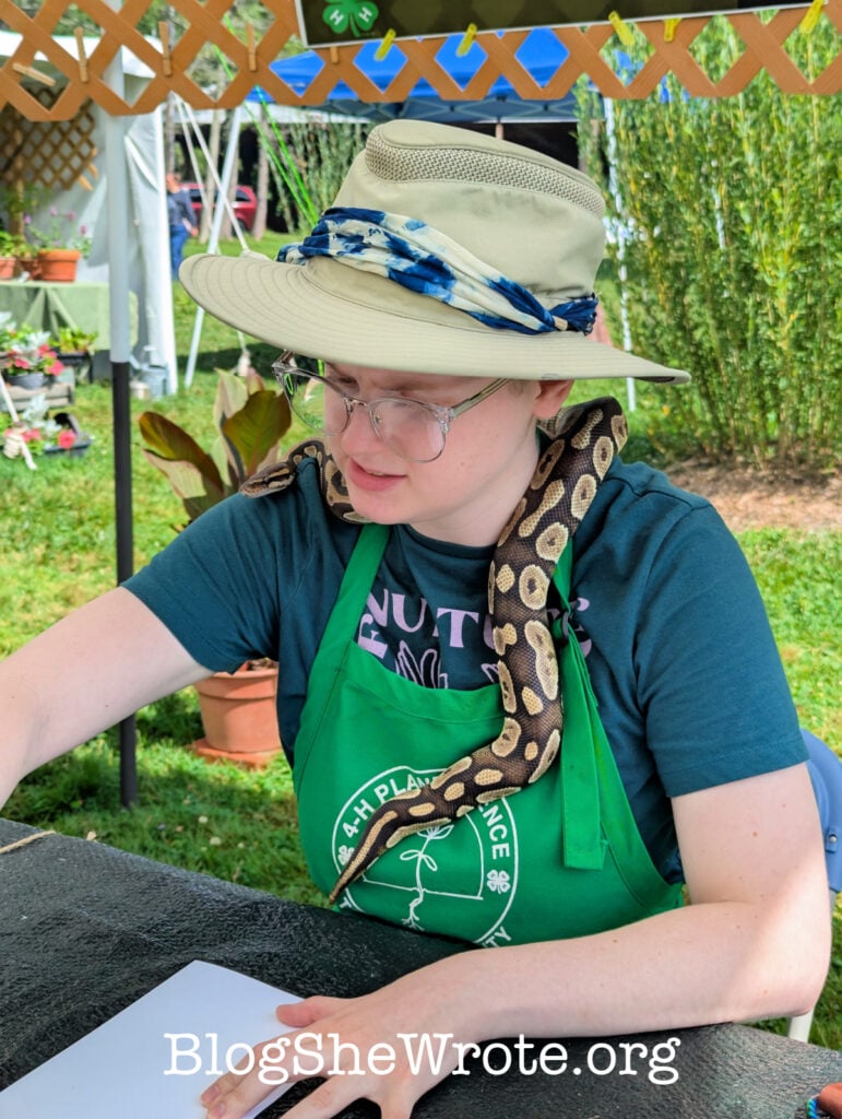 Young woman sitting and sketching with a hat on and a snake around her neck
