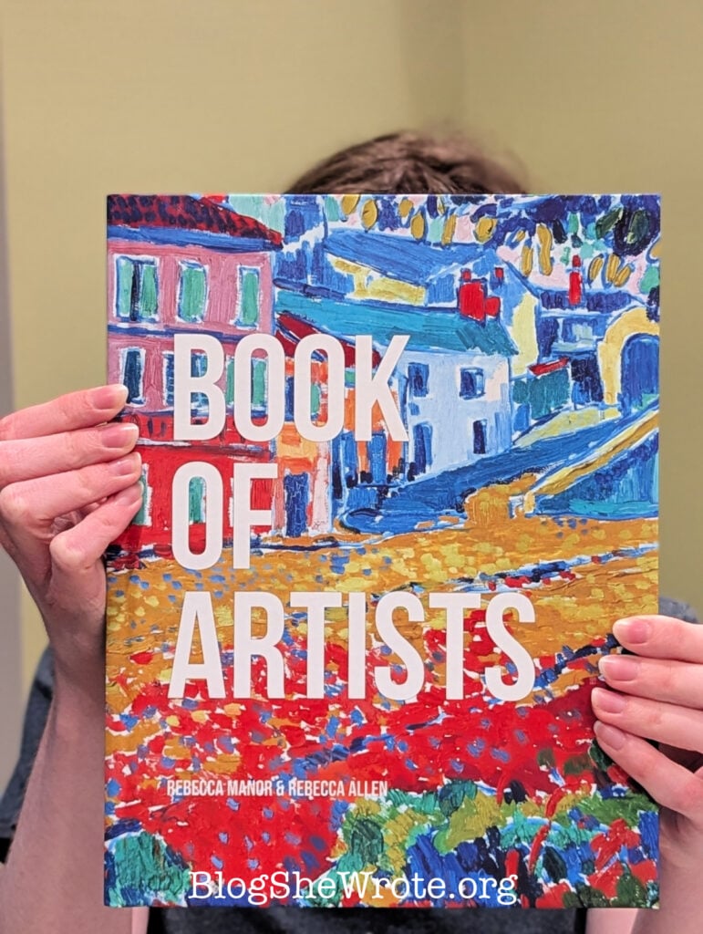 A book with the title Book of Artists being held up with two hands showing over the cover